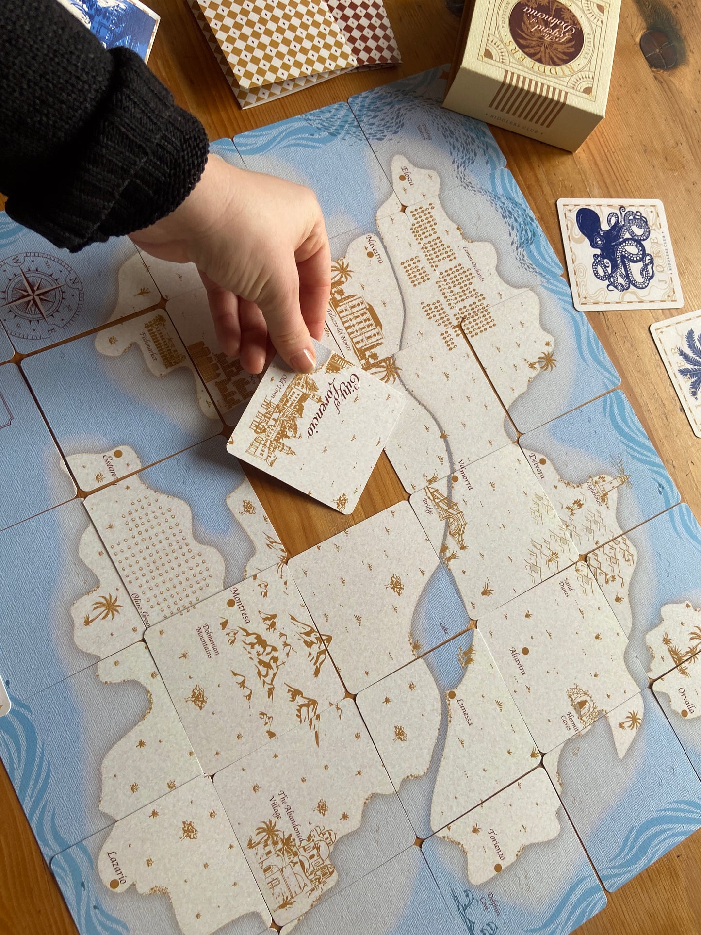 How to play our Jigsaw Island game Hand placing a card on a tiled map with decorative elements, 